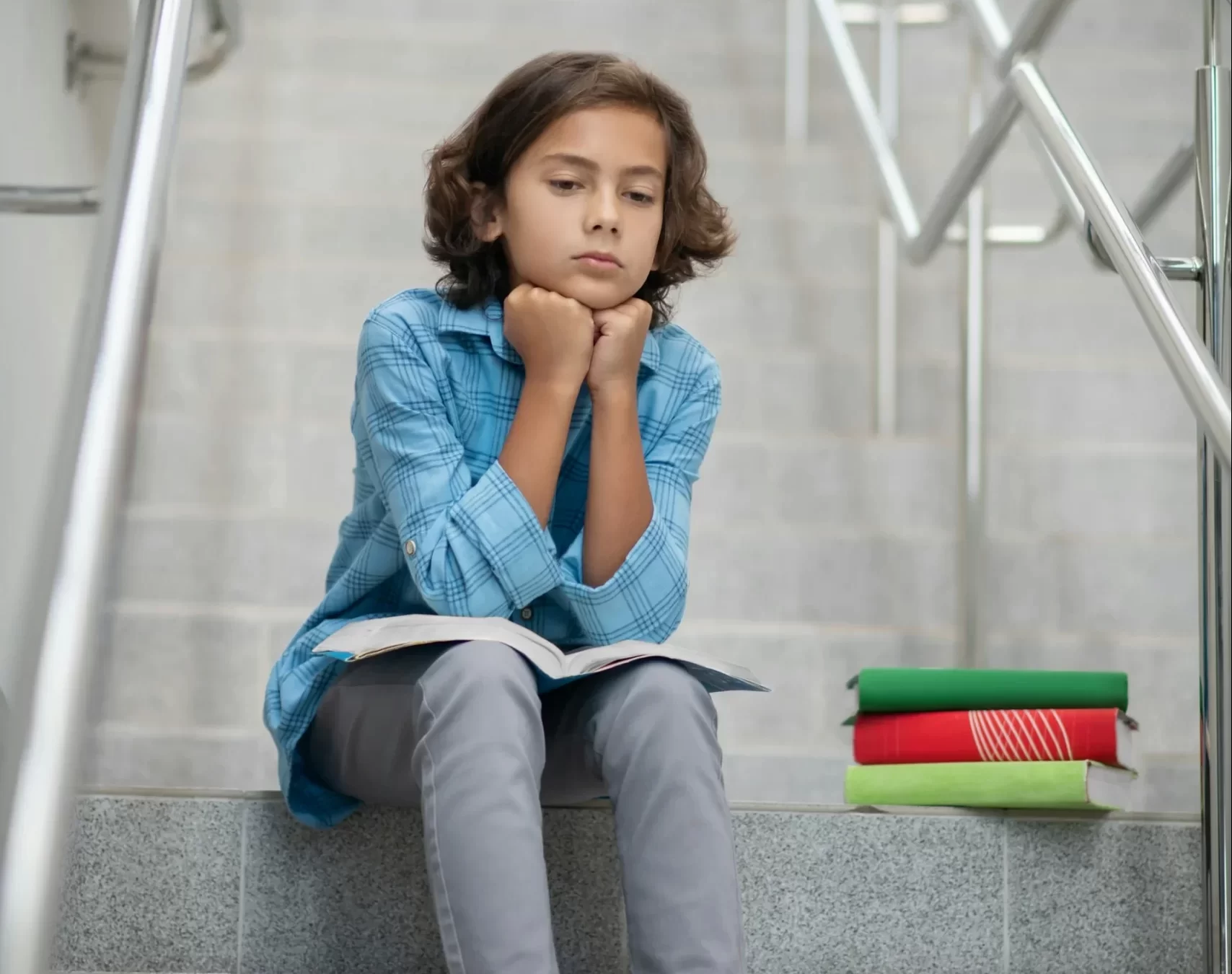 photo - a girl sitting on the stairs looking sad with a book on her knees and more books next to her waiting for her tuition online session