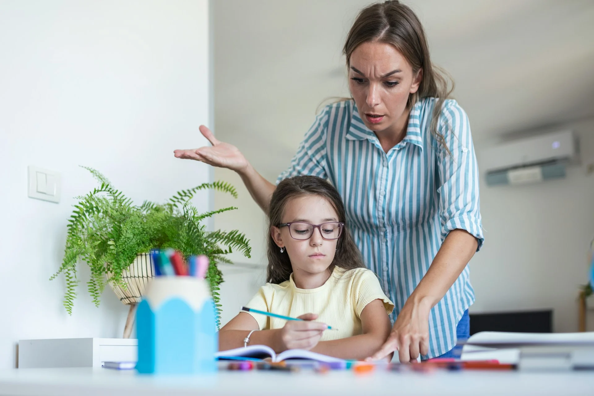 photo - a girl with verbal processing problems is sitting at a table with homework in front of her and her mother is standing behind her with a confused look on her face