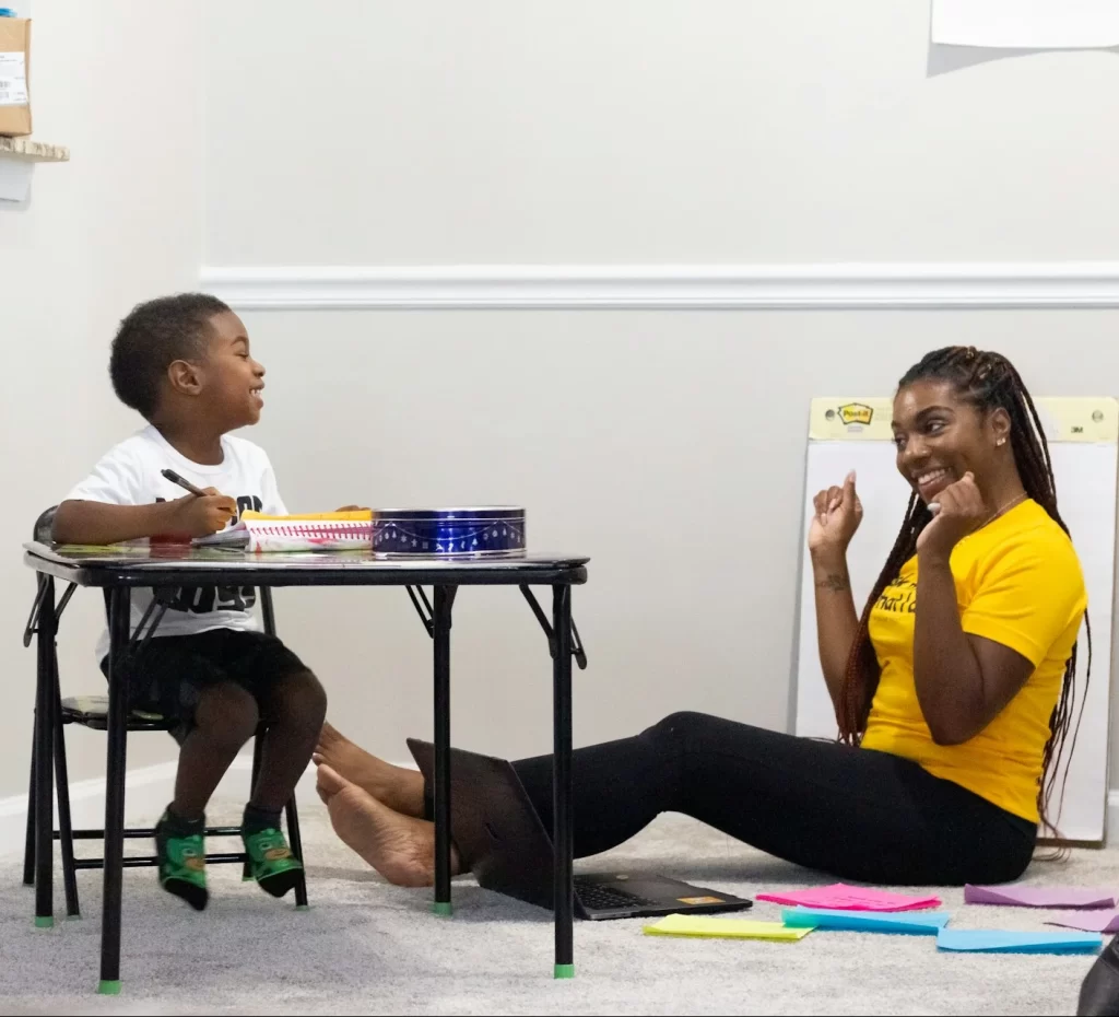 photo - Home schooling tutor sitting on the floor with a boy sitting at a desk smiling and cheering