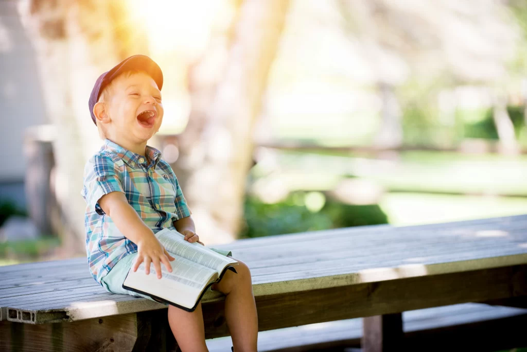 Photo - A toddler sitting on a bench, laughing while holding a book learning in special needs primary schools